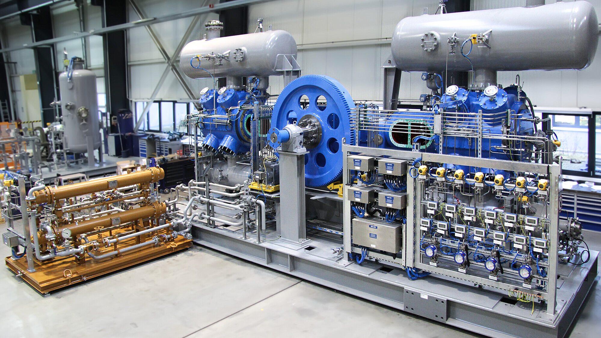 Shown is a blue 2-crank reciprocating compressor in boxer design with flywheel and pulsation tanks as well as the associated panel, installed on a skid. Next to it is the oil system.